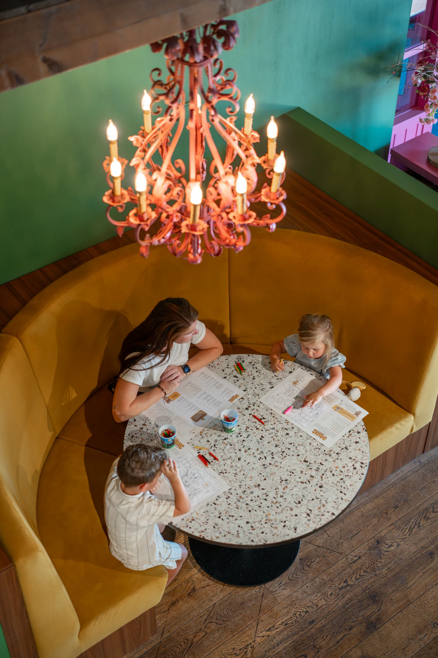Gezin kleurt aan tafel in de gele zithoek onder de roze kroonluchter bij Pluis